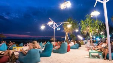 GILI AIR, INDONESIA - JULY 2, 2019: zooming out timelapse of tourists sitting in soft armchairs at brightly illuminated night beach and  enjoying fresh evening and night ocean