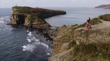 Okyanusun üzerindeki yüksek uçurumda dans eden güzel elbiseli çingene kadının hava panoramik görüntüsü.