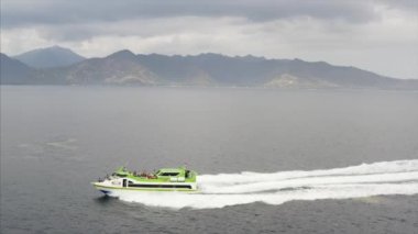 Gili Air, Endonezya - 4 Temmuz 2019: Yeşil feribotun havadan panoramik manzarası turistlerin okyanus dalgalarında hızla ilerlediği arka planda dağlarla birlikte inanılmaz bir ada  