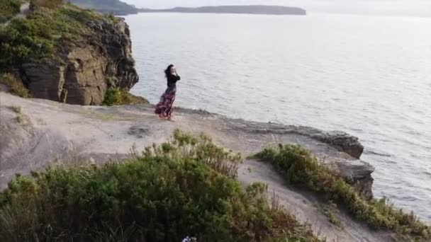 空中全景 身穿漂亮衣服 头晕目眩的女人站在高高的悬崖上俯瞰大海 — 图库视频影像