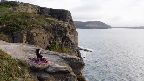空中全景 身穿漂亮衣服 头晕目眩的女人坐在高高的悬崖上俯瞰大海 — 图库视频影像