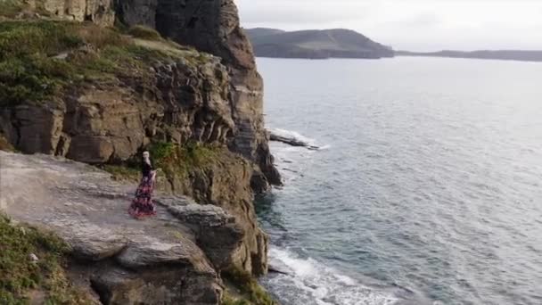 空中全景 身穿漂亮衣服 头晕目眩的女人站在高高的悬崖上俯瞰大海 — 图库视频影像
