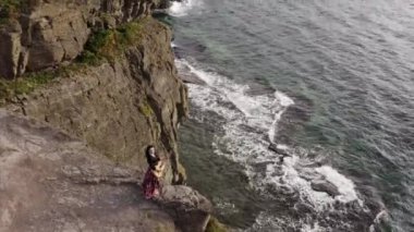 Okyanusun yukarısındaki yüksek kayalıklarda gitar çalan güzel elbiseli çingene kadının hava panoramik görüntüsü. 