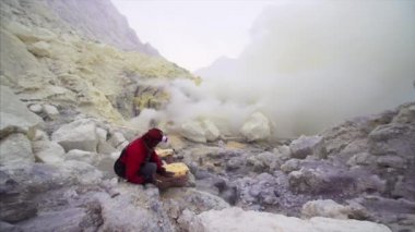 JAVA, INDONESIA - JULY 7, 2019: Indonesian man loading sulphur of Ijen volcano in baskets surrounded by poisonous fumes 