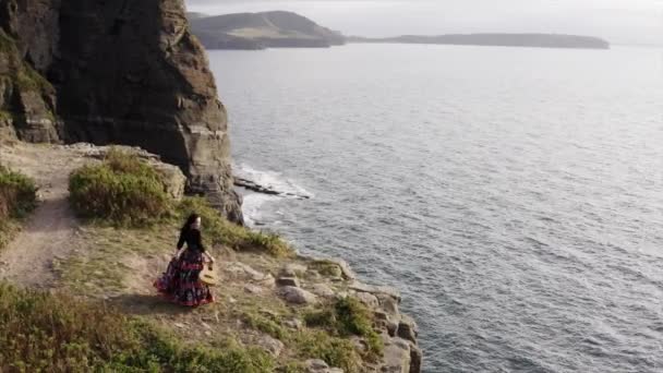 空中全景 身穿漂亮衣服 身披吉它 身披长袍 身披长袍 身披长袍 身披长袍的女子在海面之上的悬崖上翩翩起舞 — 图库视频影像