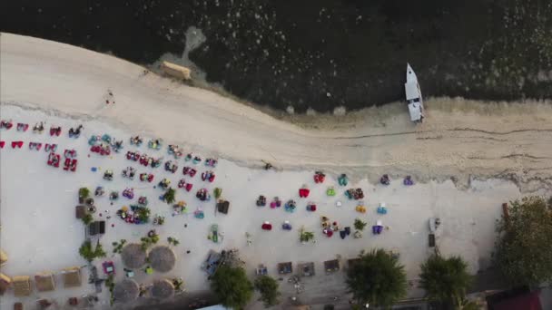 リゾートのわら傘の近くのアームチェアで新鮮な夜を楽しむ観光客と砂のビーチの空中ビュー エア島 インドネシア — ストック動画