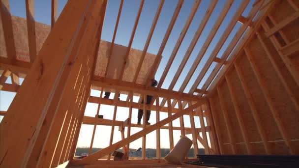 View of builders covering roof with waterproofing layer at unfinished frame house interior with wooden beams of roof and walls with blue sky on background — Stock Footage View Builders Covering Roof Waterproofing Layer Unfinished Frame House Interior Royalty Free Stock Video