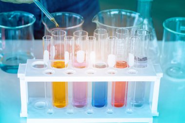 Bir bilim laboratuarında deneyciler kırmızı, turuncu, mavi ve pembe kimyasallar bırakıyor. Laboratuvardaki masada test tüpleri ve ekipmanlar bulunan test tüpüne test etmek için.