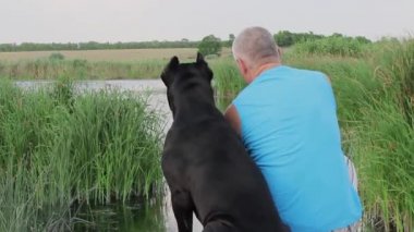 Bir yaz günü, çok sevdiği siyah Cane Corso köpeğiyle birlikte nehir kıyısında oturup akan sulara ve sazlıklara bakan genç bir adam.