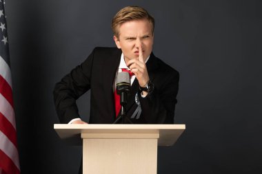 Hoşnutsuz duygusal adam siyah arka planda Amerikan bayrağıyla Tribüne sessiz jest yapıyor.