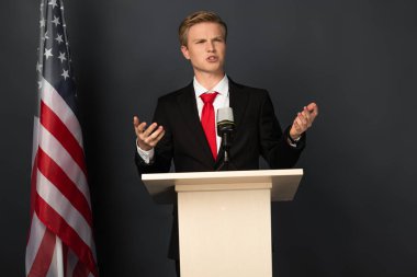 Siyah arka planda Amerikan bayrağı olan duygusal bir adam tribünde konuşuyor.