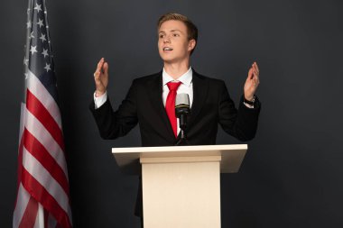 Siyah arka planda Amerikan bayrağı olan duygusal bir adam tribünde konuşuyor.