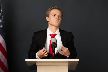 upset emotional man touching jacket on tribune with american flag on black background clipart