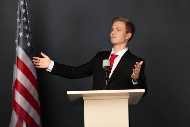 Siyah arka planda Amerikan bayrağı olan tribünde jest yapan duygusal bir adam.