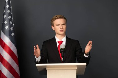 Siyah arka planda Amerikan bayrağı olan tribünde jest yapan duygusal bir adam.