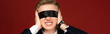man with blindfold on eyes covering ears with hands on red background clipart