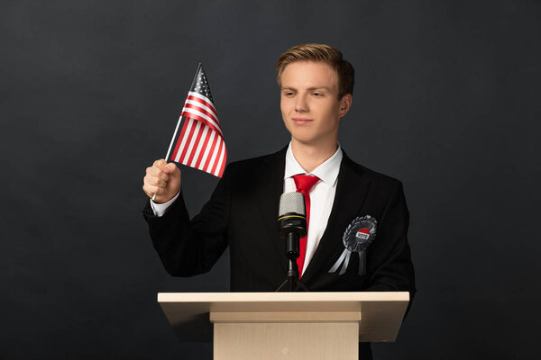 улыбающийся эмоциональный человек на трибуне с американским флагом на черном фоне
