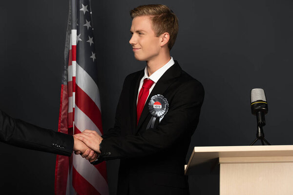 smiling emotional man shaking hand on tribune with american flag on black background