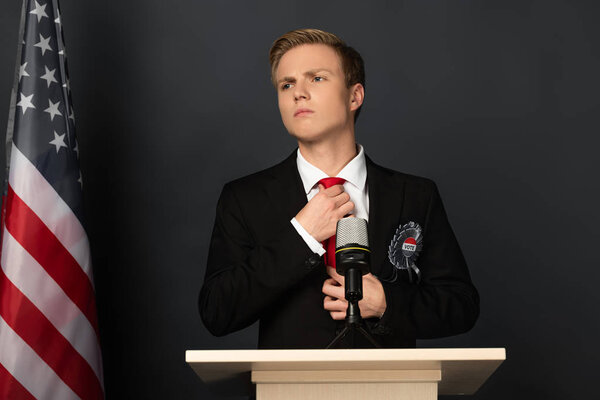 pensive man touching tie on tribune with american flag on black background