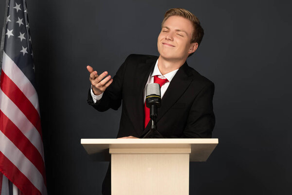 smiling emotional man gesturing on tribune with american flag on black background