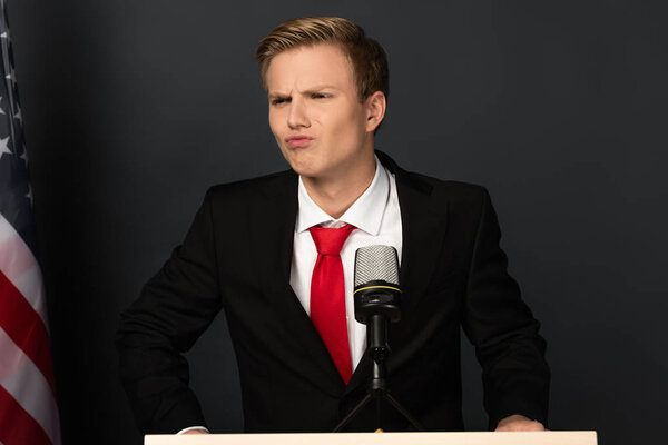 emotional man grimacing on tribune with american flag on black background