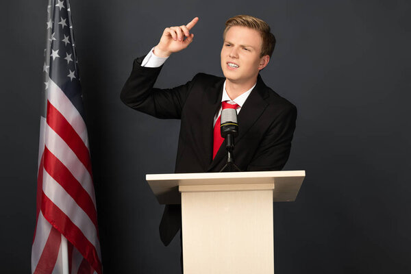 эмоциональный человек выступает на трибуне с американским флагом на черном фоне
