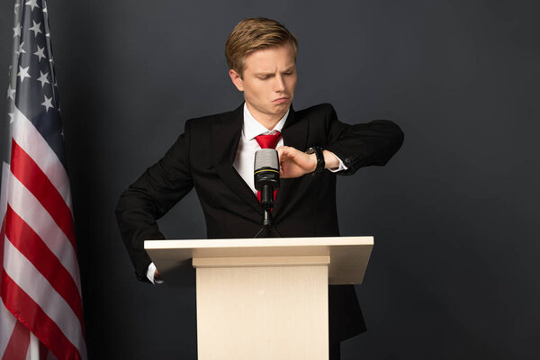эмоциональный человек смотрит на наручные часы на трибуне с американским флагом на черном фоне
