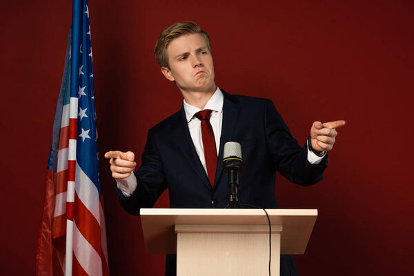 emotional man pointing with fingers on tribune near american flag on red background