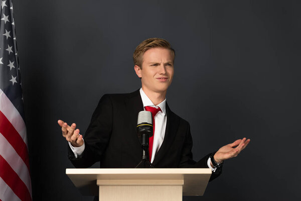 emotional man showing shrug gesture on tribune with american flag on black background