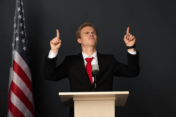 emotional man pointing with fingers upwards on tribune with american flag on black background