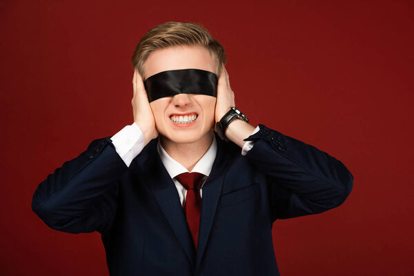 man with blindfold on eyes covering ears with hands on red background