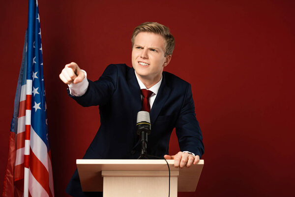 emotional man pointing with finger on tribune near american flag on red background