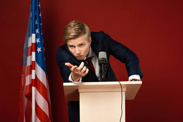 serious man pointing with finger at camera on tribune near american flag on red background
