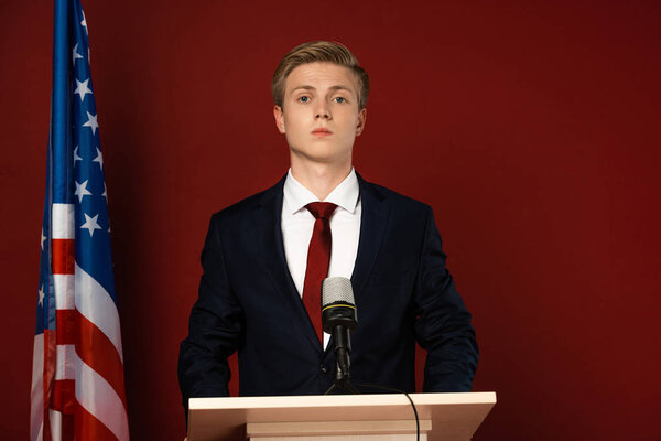 serious man on tribune near american flag on red background