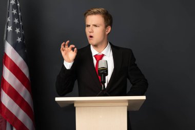 emotional man gesturing on tribune with american flag on black background clipart