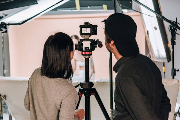 Rear view of cameraman and assistant looking at camera display while working with woman in photo studio