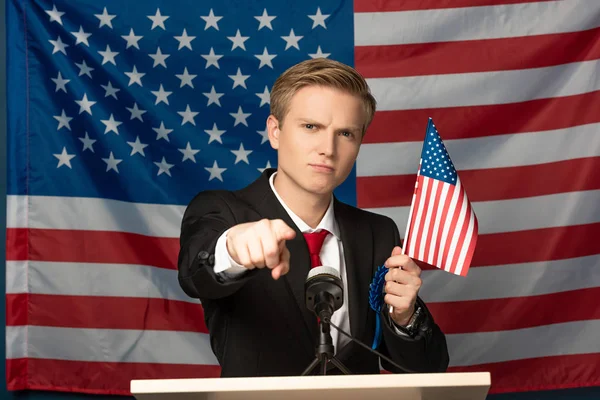 Недовольный мужчина, указывающий пальцем на камеру на трибуне на фоне американского флага — стоковое фото