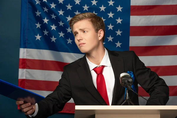 Displeased man holding clipboard on tribune on american flag background — Stock Photo