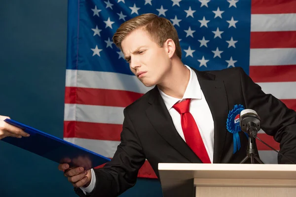 Displeased man holding clipboard on tribune on american flag background — Stock Photo