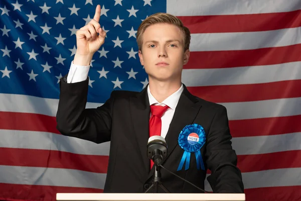 Hombre seguro de sí mismo con la mano levantada en tribuna sobre fondo de bandera americana — Stock Photo