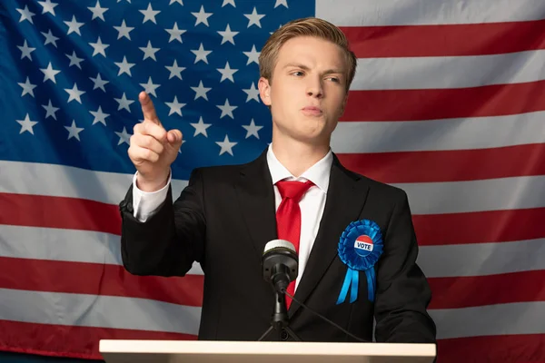 Hombre emocional señalando con el dedo en tribuna en fondo de la bandera americana - foto de stock