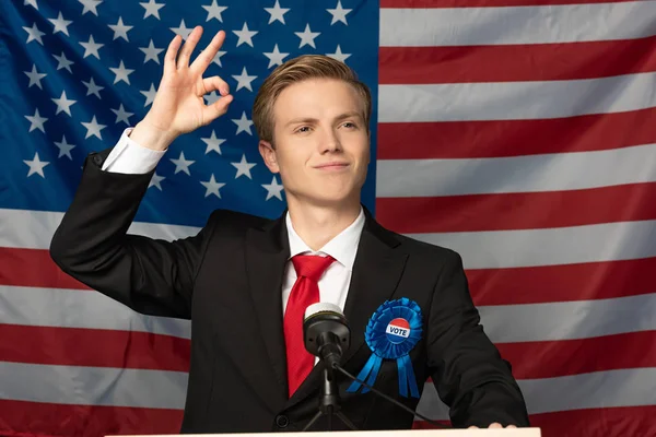 Smiling man showing ok sign while on tribune on american flag background — Stock Photo