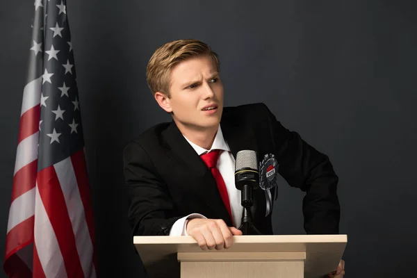 Hombre emocional indignado en tribuna con bandera americana sobre fondo negro - foto de stock