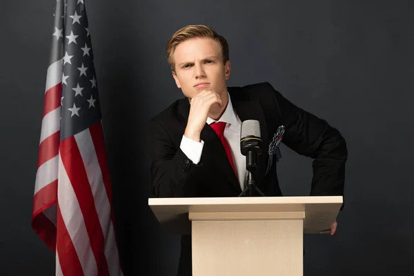 Nachdenklicher emotionaler Mann auf der Tribüne mit amerikanischer Flagge auf schwarzem Hintergrund — Stockfoto