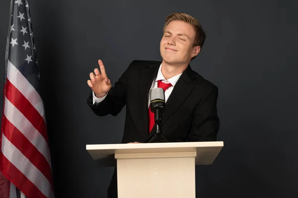 Uomo emotivo sorridente mostrando idea gesto su tribuna con bandiera americana su sfondo nero — Foto stock