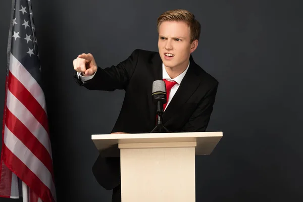 Wütender emotionaler Mann zeigt mit dem Finger auf Tribüne mit amerikanischer Flagge auf schwarzem Hintergrund — Stockfoto