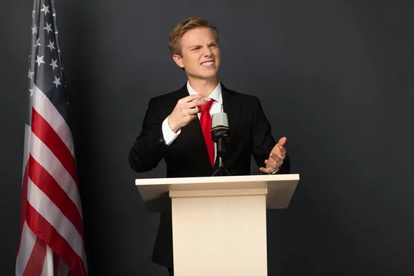 Emotional man speaking and grimacing on tribune with american flag on black background — Stock Photo