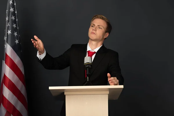 Emotional man speaking and gesturing on tribune with american flag on black background — Stock Photo