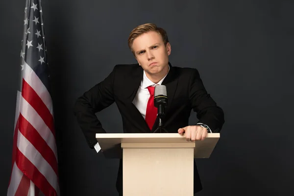 Triste hombre emocional en tribuna con bandera americana sobre fondo negro — Stock Photo