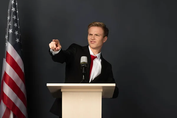 Hombre emocional señalando con el dedo a la cámara en tribuna con bandera americana sobre fondo negro - foto de stock